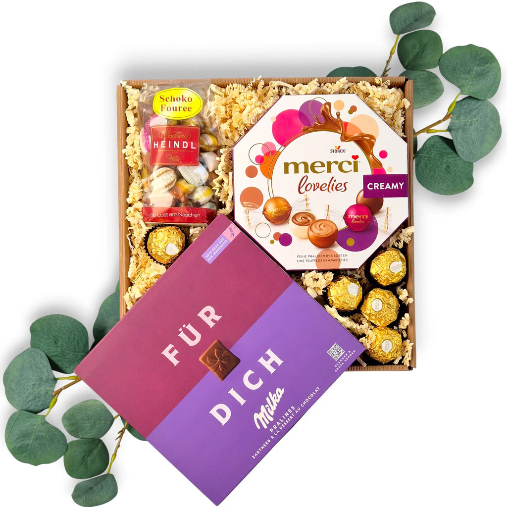 Geöffnete Geschenkbox aus braunem Naturkarton gefüllt mit Milka "Für Dich" Pralinés, einer Packung Merci Lovelies und Heindl Schoko Fouree auf hellem Füllmaterial.