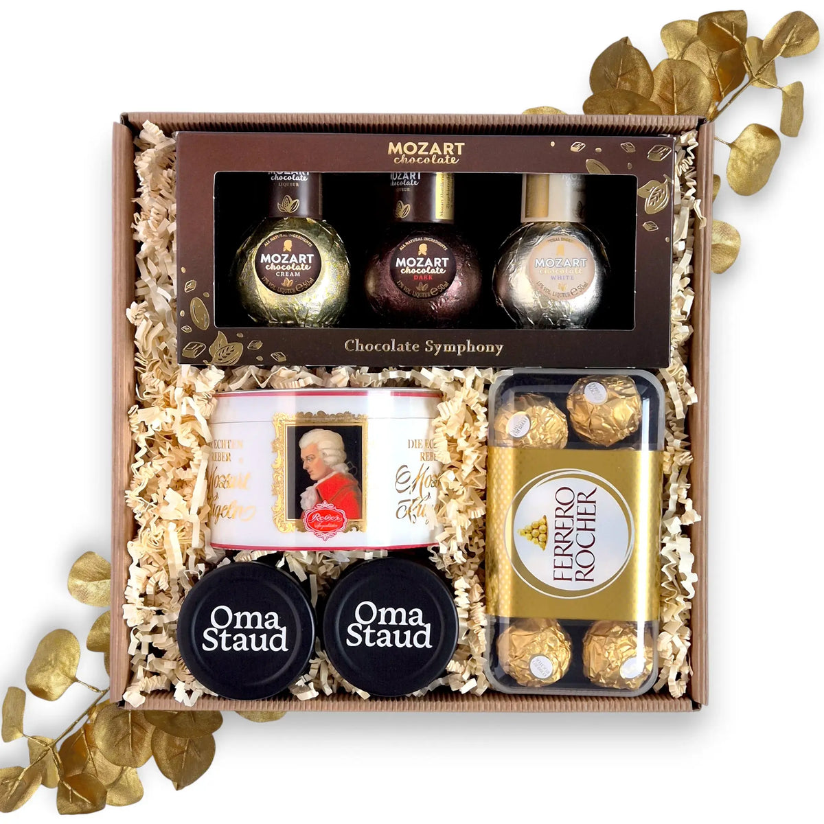 Geöffnete Geschenkbox gefüllt mit einem Mozart Schokoladenlikör Trio, einer Dose Reber Mozartkugeln, Ferrero Rocher und zwei Gläsern Staud's Konfitüre auf dunklem Füllmaterial.
