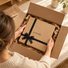 Frau packt eine geschenkify Geschenkbox mit schwarzer Schleife aus dem Versandkarton aus, sicherer Geschenkversand